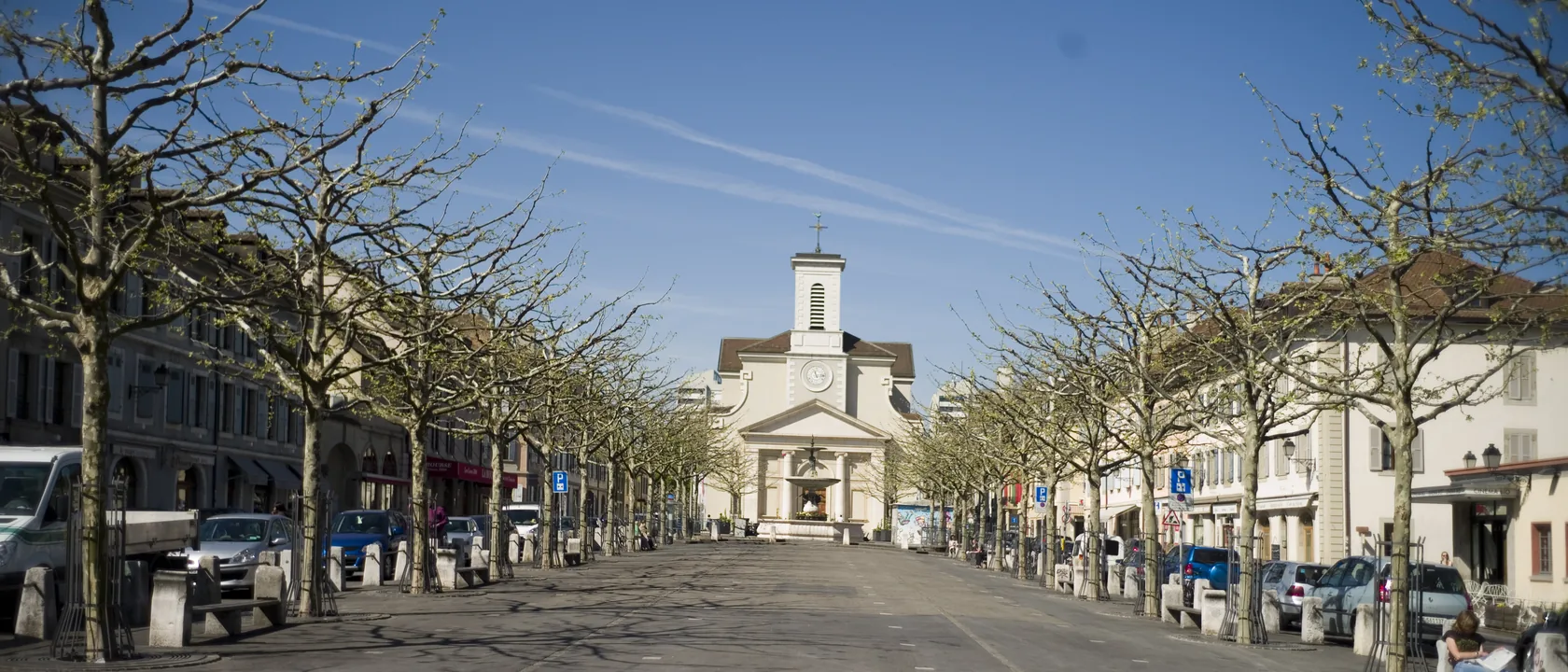 Place du Marché à Carouge bordée de platanes, avec l'église Sainte-Croix en fond et les maisons basses sardes de part et d'autre, secteur desservi par SOS Relevage